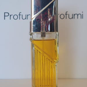 Könnte beinhalten: Eine Vintage-Parfümflasche mit silberner Kappe und einem transparenten, geriffelten Glaskörper. Die Flasche enthält eine goldfarbene Flüssigkeit. Der Text "Cliche" ist auf der Kappe sichtbar. Der Hintergrund enthält das Wort "Profumi".