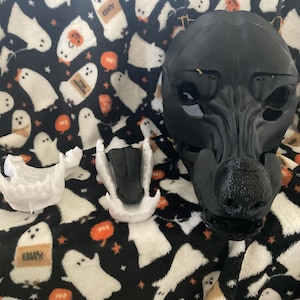 May include: A black dog mask with a textured snout and eye openings, along with two white jaw pieces and a black jaw piece. The items are displayed on a black blanket with white ghosts and orange pumpkins.