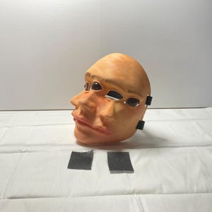 Puede incluir: Una máscara facial de color bronceado con un diseño surrealista de dos perfiles faciales superpuestos. La máscara tiene aberturas oculares oscuras y brillantes, además de rasgos esculpidos realistas que incluyen narices y labios. Dos pequeñas pestañas de tela negra están unidas al lateral.