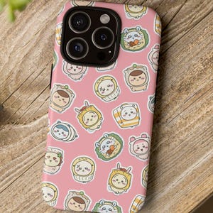 Könnte beinhalten: Eine rosa Handyhülle mit einem sich wiederholenden Muster von Cartoon-Charakteren in Kostümen. Die Hülle hat einen schwarzen Rand und einen Kameraausschnitt. Die Handyhülle liegt auf einer Holzoberfläche.