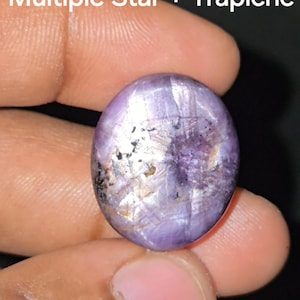 May include: A polished, oval gemstone with a star pattern and a trapiche design. The stone is mainly purple with silver and black inclusions. The text "Multiple Star + Trapiche" is visible in white.