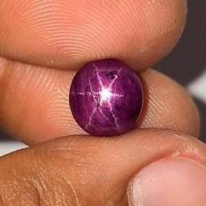 May include: A round, cabochon-cut star ruby gemstone with a deep magenta-purple hue. The stone features a distinct, centered six-rayed star pattern, known as asterism, which reflects light in a bright, sharp white cross shape across the surface.