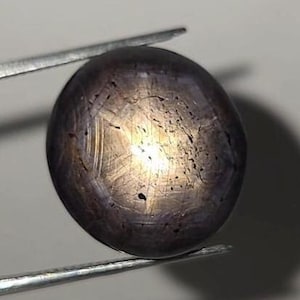 Puede incluir: Una piedra preciosa de zafiro estrella gris oscuro de forma redonda con un efecto de asterismo de seis rayos visible en el centro. La piedra tallada en cabujón muestra una textura suave y pulida con sutiles inclusiones internas, sujeta por pinzas.