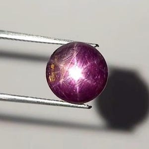 May include: A round, cabochon-cut star ruby gemstone with a deep magenta-red hue. The stone displays a distinct six-rayed star effect, known as asterism, caused by light reflecting off internal needle-like inclusions within the polished surface.