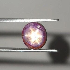Pode incluir: Uma pedra preciosa de rubi estrela de corte cabochon redondo com um efeito de estrela de seis raios distinto centrado na sua superfície. A pedra exibe um tom vermelho arroxeado profundo e é mantida entre as pontas metálicas prateadas.