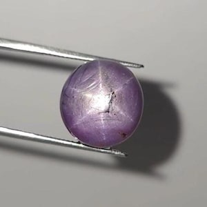 Può includere: Una gemma di zaffiro stellato a taglio cabochon rotondo con una tonalità viola chiaro traslucida. La pietra mostra un distinto effetto a stella a sei raggi sulla sua superficie lucida e bombata, tenuta da una pinzetta in metallo color argento.