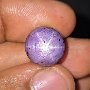Könnte beinhalten: Ein runder, im Cabochon-Schliff gehaltener Sternsaphir-Edelstein mit hellvioletter Färbung. Der Stein zeigt einen ausgeprägten sechsstrahligen Sterneffekt, bekannt als Asterismus, der auf seiner Oberfläche zentriert ist, sowie kleine, dunkle Einschlüsse.