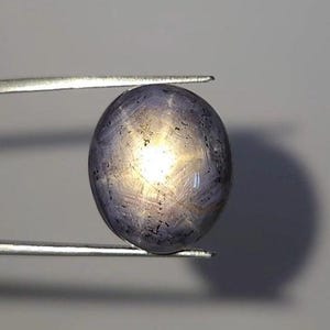 May include: A round, cabochon-cut star sapphire gemstone with a greyish-blue hue and a visible six-rayed star effect centred on its surface. The gemstone is held securely by a pair of thin, silver-toned metal tweezers against a plain background.