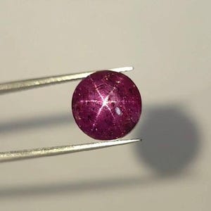 Puede incluir: Una piedra preciosa de rubí estrella redonda de color magenta intenso que presenta un patrón de estrella de seis rayos en su superficie. La piedra tallada en cabujón muestra un tono rosa rojizo vibrante y saturado, sostenida por unas pinzas metálicas finas.