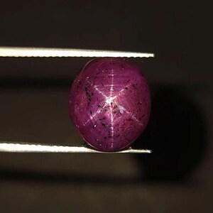 Può includere: Una gemma di rubino stellato a taglio cabochon rotondo con una tonalità rosso magenta intenso. La pietra mostra un distinto effetto di asterismo a sei raggi centrato sulla superficie, caratterizzato da linee di luce che si incrociano su fondo.
