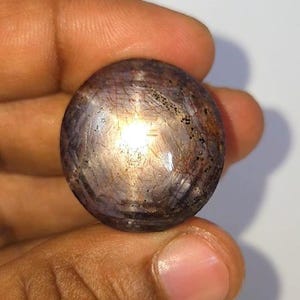 Op de afbeelding: Een ronde, gepolijste edelsteen met een sterachtig effect. De steen heeft een donkerpaarse tint met gouden en zilveren interne reflecties. De edelsteen wordt in een hand gehouden tegen een witte achtergrond, wat de kleur en textuur benadrukt.