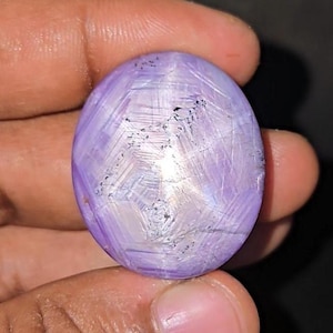 May include: An oval-shaped cabochon gemstone with a smooth, polished surface. The stone displays a light purple colour with a distinct, intersecting white grid-like pattern across its face, featuring small dark inclusions throughout the structure.