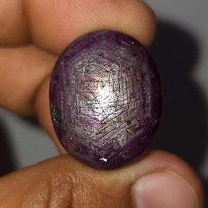 Könnte beinhalten: Ein ovaler Sternrubin-Edelstein im Cabochon-Schliff mit einem tiefen rot-violetten Farbton. Die Oberfläche zeigt einen ausgeprägten sechsstrahligen Asterismus-Effekt, der durch sich kreuzende Linien ein Sternmuster in der Mitte bildet.