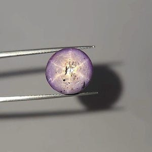 Könnte beinhalten: Ein runder, polierter, lila Edelstein mit sternförmigem Muster, gehalten von einer silbernen Pinzette. Der Edelstein hat ein gesprenkeltes Aussehen und ein zentrales Sterndesign. Der Hintergrund ist einfarbig grau.