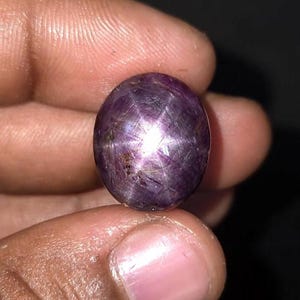 Könnte beinhalten: Ein polierter, ovaler Sternrubin-Edelstein mit einem tiefen violett-roten Farbton und einem deutlichen sechsstrahligen Sternmuster auf seiner glatten, im Cabochon-Schliff bearbeiteten Oberfläche. Der Stein weist natürliche Einschlüsse und einen hellen Lichtreflex auf.
