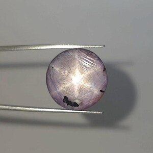 以下が含まれることがあります： 銀色のピンセットで保持された、星模様の丸いラベンダー色の宝石。 研磨された宝石は、中心から放射状に広がる六芒星のデザインが特徴です。 表面には暗いインクルージョンが見られます。