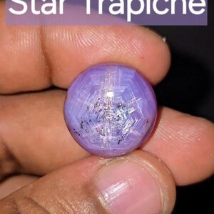 Peut inclure: Une pierre précieuse sphérique violette et facettée, avec un motif en étoile. La gemme est tenue entre les doigts, avec le texte "Star Trapiche" dans un rectangle violet au-dessus.