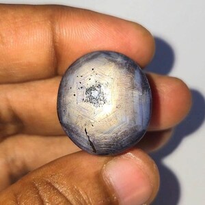 Può includere: Una gemma di zaffiro stellato a taglio cabochon rotondo, caratterizzata da una tonalità blu grigiastro con un evidente effetto a stella a sei raggi centrato sulla superficie. La pietra presenta una finitura liscia e lucida con inclusioni.