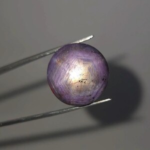 Puede incluir: Una gema redonda, de color rosa púrpura, sujeta con pinzas. La gema tiene un patrón en forma de estrella en su superficie, con un centro más claro, casi dorado. El fondo es gris claro liso.