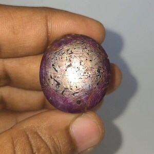 Op de afbeelding: Een bolvormige, paarse edelsteen met een metallic, goudkleurige glans. Het oppervlak heeft donkere, onregelmatige markeringen. De edelsteen wordt in een hand gehouden tegen een lichtgrijze achtergrond. Het object heeft een diameter van ongeveer 2,5 cm.