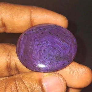May include: A polished, round, purple gemstone with a distinctive star-like pattern. The stone is held in a hand, highlighting its colour and intricate design. The gemstone's surface has a glossy finish and dark inclusions.