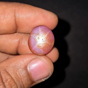 May include: Oval-shaped, star ruby gemstone with a pinkish-red hue and a prominent star effect. The gem has a polished surface and displays a play of light. The background is dark, emphasizing the gem's color and brilliance.