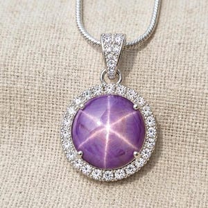May include: A silver-toned necklace featuring a round, purple star sapphire cabochon pendant. The centre stone displays a distinct six-rayed star pattern and is surrounded by a halo of small, clear, sparkling stones set in a silver-coloured metal frame.