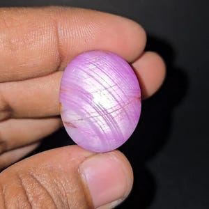 Puede incluir: Gema ovalada y pulida con un patrón arremolinado y rayado en tonos rosa y blanco. La gema se sostiene entre los dedos sobre un fondo oscuro. La piedra tiene una superficie pulida y reflectante.