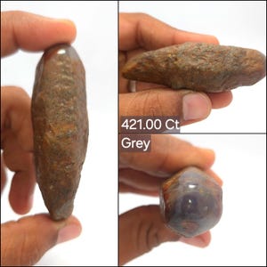 Könnte beinhalten: Ein länglicher Stein mit rauer Textur, brauner, verwitterter Oberfläche und einem glatten, polierten grau-violetten Ende. Der Stein ist mit 421.00 Ct und Grey beschriftet. Die Oberfläche zeigt natürliche mineralische Variationen und einen deutlichen Kontrast zwischen den rohen und polierten Bereichen.