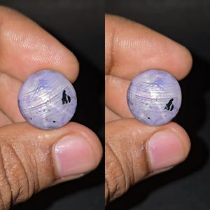 Op de afbeelding: Een ronde, lichtpaarse edelsteen met een gepolijst oppervlak. De steen heeft een wervelend, gestreept patroon met donkere insluitsels. De edelsteen wordt tussen twee vingers tegen een donkere achtergrond gehouden.