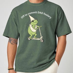 May include: Green t-shirt with the text "Off to commit DAD foolery." The shirt features a cartoon frog riding a scooter and wearing a wizard hat. The word "Moss" is in the upper left corner.