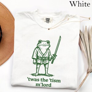 May include: White t-shirt with a green illustration of a frog in a kimono holding a sword. The text "'twas the 'tism m'lord" is printed below the frog. The shirt has a crew neck and short sleeves.