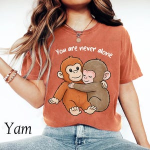 Puede incluir: Camiseta naranja quemado con una ilustración de dibujos animados de dos monos abrazándose y el texto "You are never alone". La camiseta está hecha de un material suave.