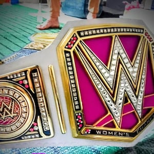 May include: A championship belt with a white strap and gold accents. The center plate features a large, sparkling "W" over a pink background. The words "WOMEN'S CHAMPION" are visible.