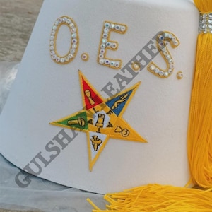 Puede incluir: Sombrero de fieltro blanco con las letras "O.E.S." en oro y pedrería. Un emblema de estrella colorida está bordado en la parte delantera. Una borla amarilla cuelga del lateral. El sombrero es un símbolo de la Orden de la Estrella del Este.