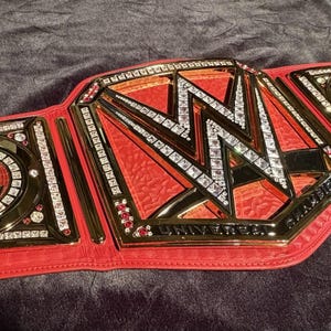 Puede incluir: Un cinturón de campeonato de cuero rojo con detalles dorados y el logotipo de la WWE. La placa central presenta las letras "W" y "W" con un patrón de diamantes. El cinturón tiene un borde dorado y está tachonado con pedrería.
