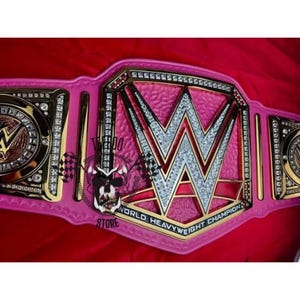 May include: A pink and gold championship belt with the WWE logo in the center. The belt has a pink strap with gold accents and is encrusted with rhinestones. The words "World Heavyweight Champion" are visible.