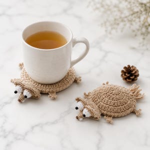 May include: Two round, tan crocheted coasters shaped like squirrels with white and black bead eyes, small ears, and bushy tails. One coaster supports a white ceramic mug filled with light brown liquid, while the other lies flat nearby.