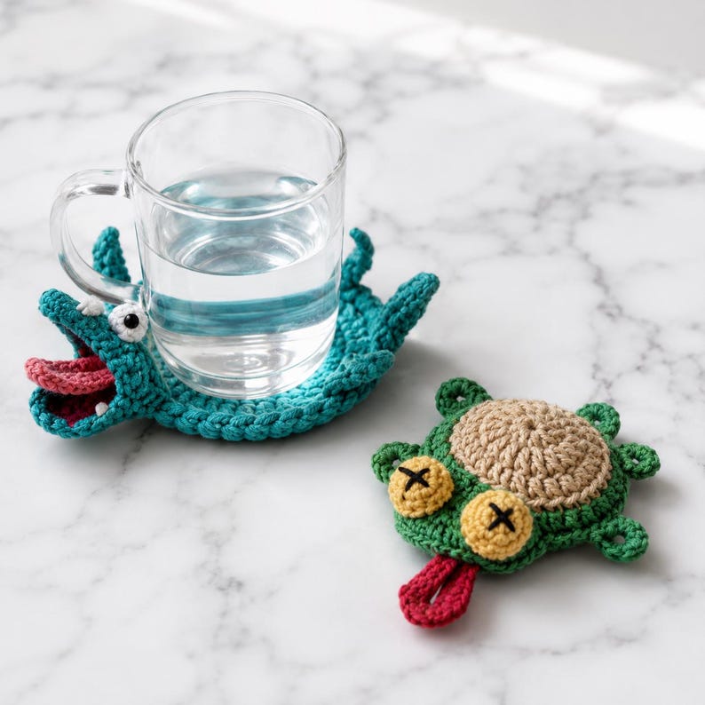 May include: Two handmade crochet coasters shaped like cartoon creatures. One teal coaster features an open mouth with a pink tongue and white teeth, supporting a clear glass mug. The second coaster is green with a tan center and yellow eyes.