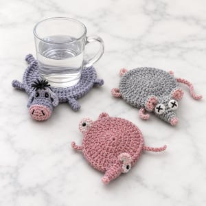 May include: Three round crochet coasters shaped like animals, including a purple donkey, a gray mouse with X-shaped eyes, and a pink pig. A clear glass mug filled with water sits on top of the purple donkey coaster, which features a snout and ears.