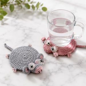 May include: Two round, crocheted animal coasters, one in light gray and one in dusty pink. Each coaster features a flat, circular body with a small head, ears, tiny limbs, and a tail. The eyes are white circles with black stitched X marks.