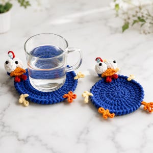 May include: Two round, dark blue crocheted coasters shaped like chickens. Each coaster features a white head with black eyes, an orange beak, a red wattle, and small yellow feet extending from the sides. A clear glass mug sits on one coaster.