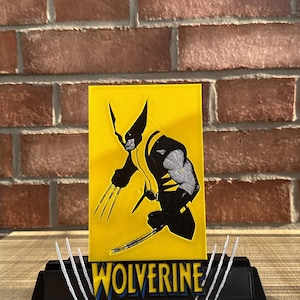 May include: A rectangular yellow plaque featuring a black and silver graphic of the character Wolverine with extended claws. The plaque sits on a black base with the word WOLVERINE in yellow letters, flanked by three white claw marks on each side.