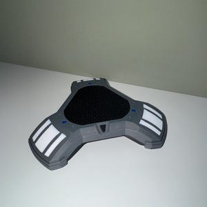 Peut inclure: Un appareil triangulaire gris avec un centre en maille noire et des accents blancs et gris. L'appareil comporte trois extensions arrondies avec des rayures blanches et de petits d&eacute;tails bleus. Il repose sur une surface blanche.