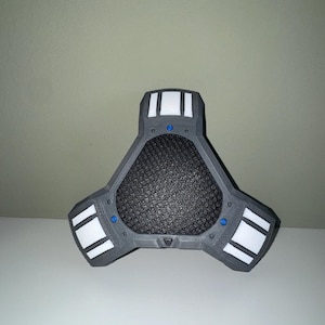 Peut inclure: Un appareil triangulaire gris avec un centre en maille noire et des accents rectangulaires blancs. De petits cercles bleus sont visibles sur l'appareil. L'objet est sur une surface blanche.