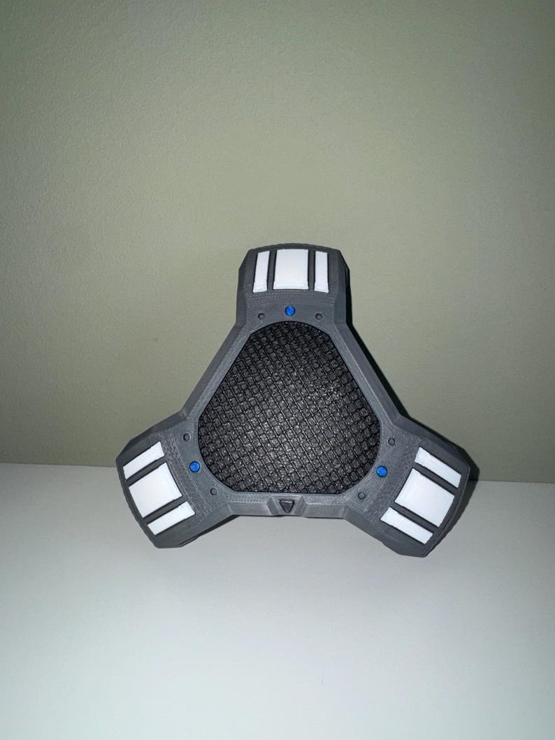 Peut inclure: Un appareil triangulaire gris avec un centre en maille noire et des accents rectangulaires blancs. De petits cercles bleus sont visibles sur l'appareil. L'objet est sur une surface blanche.