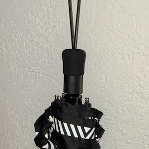 Op de afbeelding: Een zwarte inklapbare paraplu met een wit geometrisch patroon hangend aan een zwarte muurhaak. De paraplu heeft een zwart handvat en een dun zwart bandje dat over de haak met witte punt loopt, waardoor de gevouwen stof vast blijft zitten.