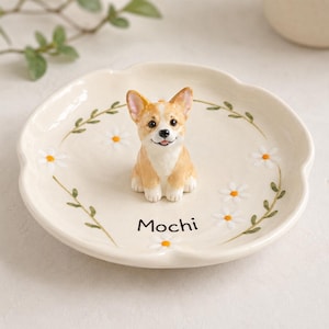 May include: A cream-colored ceramic trinket dish featuring a three-dimensional figurine of a tan and white Corgi dog in the center. The dish is decorated with painted white daisy flowers and green vines, with the word Mochi printed in black.