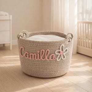 Puede incluir: Una cesta de almacenamiento tejida beige con el nombre "Camilla" en escritura rosa y una aplicación de flor blanca y rosa. La cesta tiene dos asas y está rellena con una manta blanca. Ideal para la decoración de la guardería.