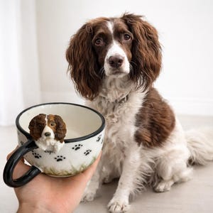 May include: A white ceramic mug featuring a black rim and a three-dimensional sculpted brown and white spaniel dog head attached to the side. The exterior of the mug is decorated with black paw prints and green grass patterns, with the word Cupid visible.